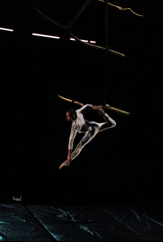 hombre acrobáta vestido de blanco estirandose suspendido en un palo de madera acrobacia